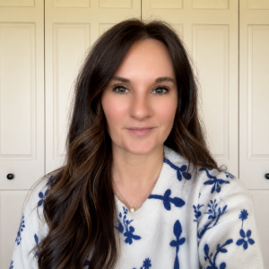 A woman with long brown hair, wearing a white sweater with blue floral patterns, smiles softly at the camera. She is indoors, in front of cream-colored paneled doors.